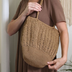 May include: A tan crochet tote bag with a rounded bottom and vertical textured pattern. The bag has a matching strap and is held by a person wearing a brown dress. The bag appears to be handmade.