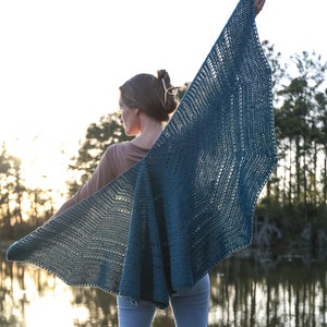 May include: A teal crochet shawl with a lacy pattern, worn by a person standing on a wooden dock by a lake.