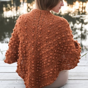 Puede incluir: Un chal de ganchillo marrón con un patrón de punto de relieve con textura. El chal está drapeado sobre los hombros de una persona y está sentado sobre una superficie de madera.