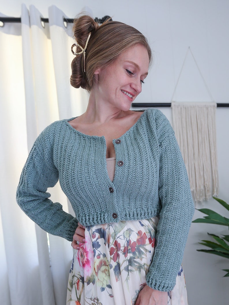 May include: A light blue crocheted cardigan with long sleeves and a button closure. The cardigan is cropped and has a ribbed texture.