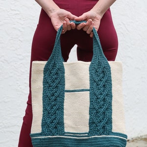 Puede incluir: Un bolso de mano de crochet de color verde azulado y blanco. El bolso tiene una base ancha y dos asas de color verde azulado.