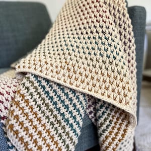 Op de afbeelding: Een gehaakt deken met een gestreept patroon in tinten bruin, groen en crème. Het deken is over een stoel gelegd.