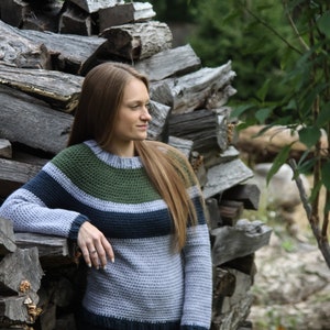 May include: A woman wearing a striped jumper with green, blue and grey stripes. She is standing next to a pile of stacked firewood.