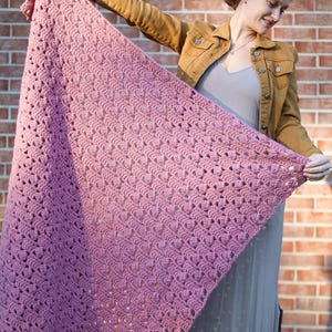 May include: A pink crocheted shawl with a delicate stitch pattern. The shawl is held by a person wearing a brown jacket and a gray dress.