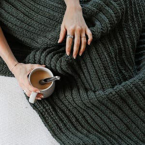 May include: A dark green, chunky knitted blanket with a ribbed texture. A white mug with a beverage and a spoon rests on the blanket. The blanket is folded, and the image is taken from an overhead perspective.