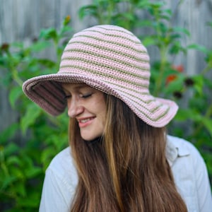 May include: A pink and green striped crochet sun hat with a wide brim. The hat is handmade and has a rounded crown. The hat is worn by a person with long brown hair and a white shirt.
