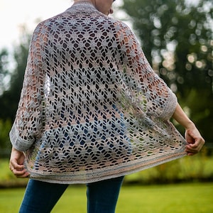 Puede incluir: Un cárdigan de crochet de tejido abierto en tonos grises, beige y melocotón. La prenda tiene un patrón texturizado y calado y mangas tres cuartos. El cárdigan se lleva sobre una camiseta oscura y vaqueros azules, con un fondo verde.