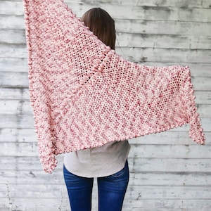 May include: A pink and white crocheted shawl with a textured bobble stitch. The shawl is draped over a person's shoulders and is flowing in the wind.