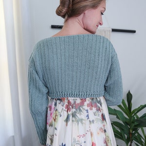 May include: A light blue crocheted cropped sweater with long sleeves. The sweater is worn over a white floral dress.