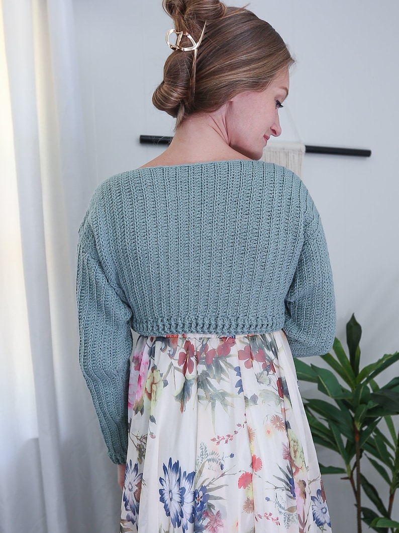 May include: A light blue crocheted cropped sweater with long sleeves. The sweater is worn over a white floral dress.