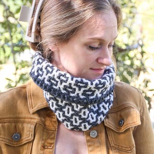 May include: A woman wearing a handmade, geometric-patterned cowl scarf in shades of grey and white. She is also wearing a brown denim jacket and has a hair clip in her hair. The scarf appears to be crocheted or knitted.