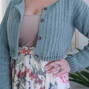 May include: A light blue crocheted cardigan with long sleeves and a button closure. The cardigan is cropped and has a ribbed texture.