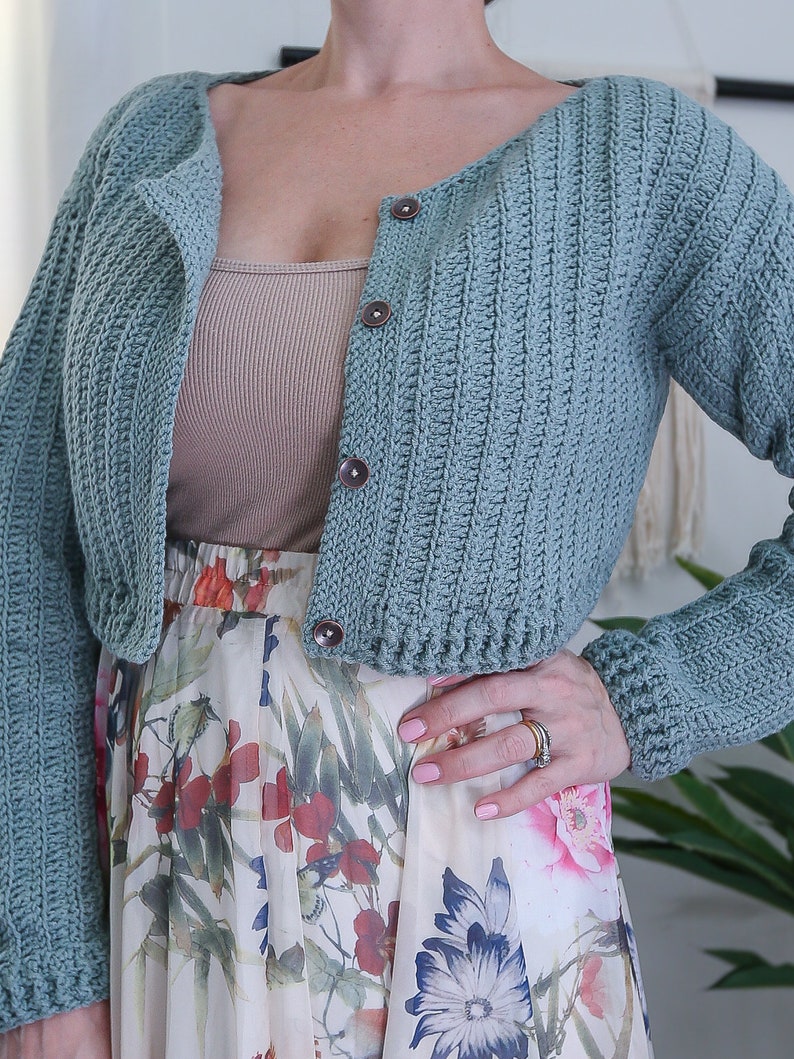 May include: A light blue crocheted cardigan with long sleeves and a button closure. The cardigan is cropped and has a ribbed texture.