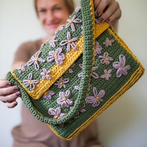 Könnte beinhalten: Eine grüne gehäkelte Tasche mit gelbem und violettem Blumendesign. Die Tasche hat einen langen Riemen und wird von einer Person gehalten.