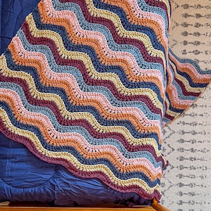 May include: A handmade crochet blanket with a wavy pattern in shades of blue, pink, orange, yellow, and burgundy. The blanket is draped over a bed, showcasing its colorful design and texture. The blanket's edge has a scalloped finish.