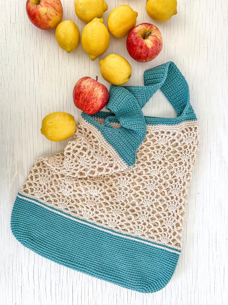 May include: A crocheted tote bag with a teal blue bottom and a beige top. The bag is made of a delicate, open-weave pattern. The bag is surrounded by lemons and apples.