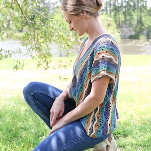 Pu&ograve; includere: Una donna indossa un top all'uncinetto a righe colorate con jeans blu e sandali. &Egrave; seduta sull'erba con la testa bassa.