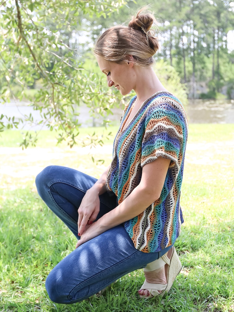 Pu&ograve; includere: Una donna indossa un top all'uncinetto a righe colorate con jeans blu e sandali. &Egrave; seduta sull'erba con la testa bassa.