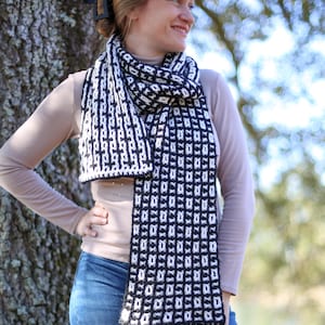 May include: A black and white crocheted scarf with a geometric pattern. The scarf is worn by a person standing in front of a tree.