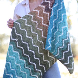 May include: A handmade crochet shawl with a chevron pattern. The shawl features a gradient of colors, transitioning from brown to green to teal and blue. White thread outlines the zig-zag design. The shawl is held up, showcasing its size and texture.