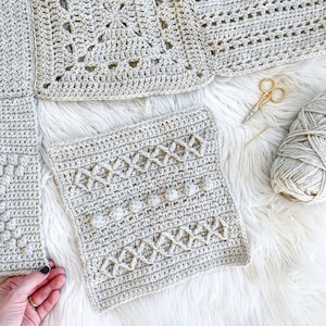 May include: A close-up of a crocheted blanket featuring a variety of stitch patterns. The blanket is made in a light beige colour and is laid out on a white fluffy surface. There are also some crochet supplies visible in the image, including a pair of scissors, a crochet hook, and a ball of yarn.