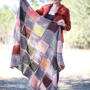 May include: A handmade, multi-colored crochet blanket featuring a diamond pattern. The blanket incorporates various shades of brown, pink, green, and yellow. The texture of the blanket is visible, showcasing the intricate crochet work. The blanket is held up, displaying its size and design.
