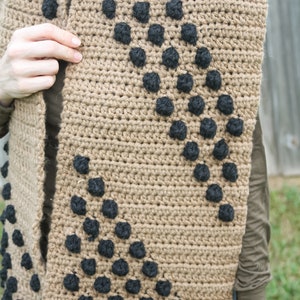 May include: A brown crocheted scarf with black yarn accents. The scarf has a diagonal pattern of black circles.