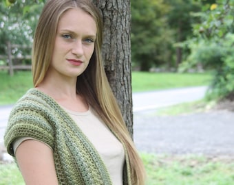 May include: A woman with long blonde hair is wearing a green crocheted cardigan over a beige tank top. She is standing next to a tree.