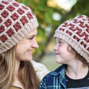 May include: Two matching crochet beanies. The beanies have a tan base with a repeating pattern of maroon squares. One beanie is worn by an adult, the other by a child. The adult is wearing a plaid shirt, and the child is wearing a blue plaid shirt.