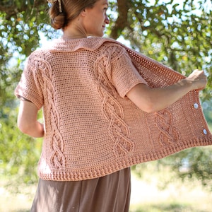 Puede incluir: Un cárdigan de crochet rosa con un patrón de trenza y mangas cortas. El cárdigan tiene un cuello y está abierto en la parte delantera.