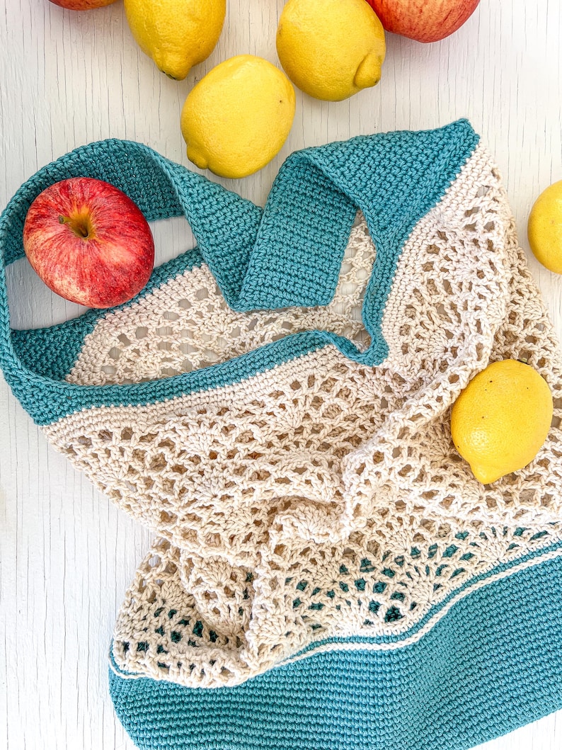 May include: A crocheted market bag with teal and beige stripes. The bag is made of a mesh material and is lying on a white surface with apples and lemons around it.