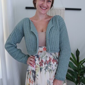 May include: A light blue crocheted cropped cardigan with long sleeves. The cardigan is open in the front and has a V-neck. The model is wearing the cardigan over a white tank top and a floral skirt.