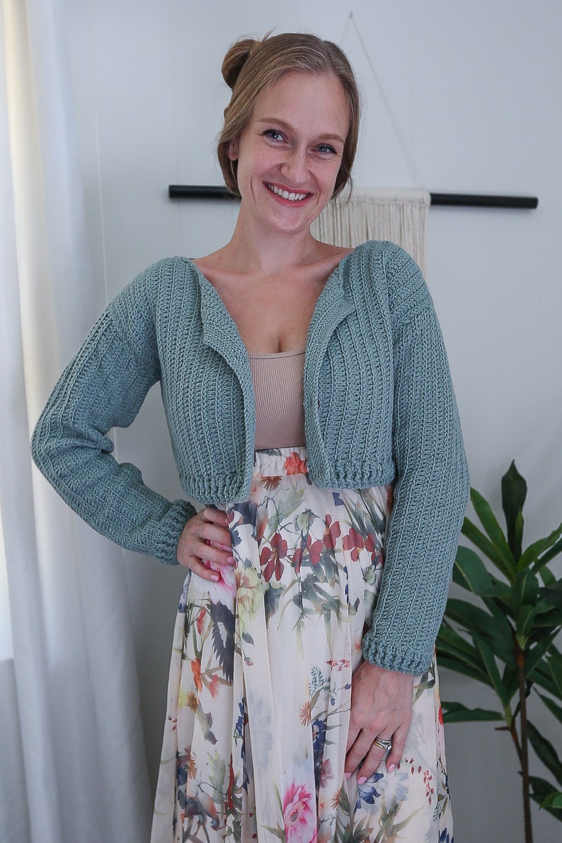 May include: A light blue crocheted cropped cardigan with long sleeves. The cardigan is open in the front and has a V-neck. The model is wearing the cardigan over a white tank top and a floral skirt.