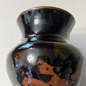 May include: A small, handcrafted ceramic vase with a glossy, dark brown glaze. The vase has a rounded body and a slightly flared rim. The surface features areas of exposed, reddish-brown clay, adding a rustic touch. The vase is approximately 4 inches tall.