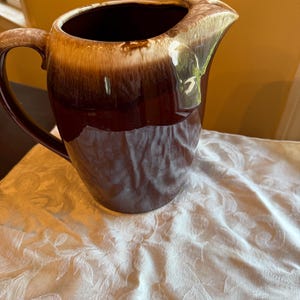 May include: A ceramic pitcher featuring a deep brown base that transitions into a lighter, creamy beige glaze around the rim and handle. The vessel has a curved spout and a single side handle, with a smooth, glossy finish throughout the exterior.