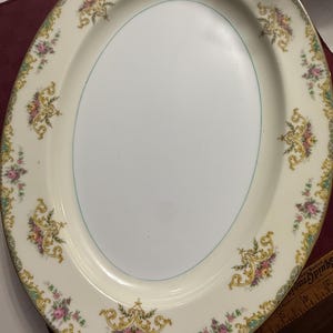 May include: An oval serving platter with a cream-colored background and a light blue inner border. The rim is decorated with floral and gold scrollwork designs. The platter has a gold trim.