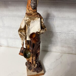 May include: A handmade paper mache figurine of a person wearing a brown and orange headscarf and holding a brown pot. The figurine is standing on a wooden base.