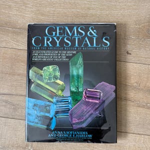 May include: A book titled "Gems & Crystals" from the American Museum of Natural History. The cover features a dark background with images of colorful gemstones and crystals. The book is an illustrated guide to the history, lore, and properties of gems and minerals.