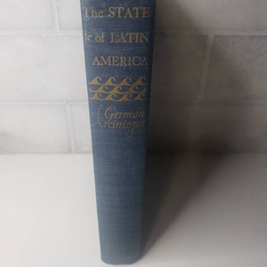 May include: A blue hardcover book with gold lettering on the spine. The title is "The State of Latin America" with a stylized wave design and the author's name "German Arciniegas".