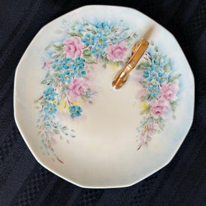 May include: A white porcelain dish with an octagonal shape, featuring a hand-painted floral pattern of pink roses and blue forget-me-nots. A gold-toned metal handle is attached to the upper right side of the dish, providing a functional grip.
