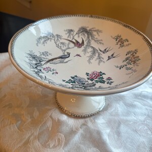 May include: A cream-colored ceramic pedestal bowl featuring a detailed transferware pattern of birds, flowering branches, and foliage in shades of pink, blue, and grey. The rim and base are accented with a delicate, dark grey beaded edge design.