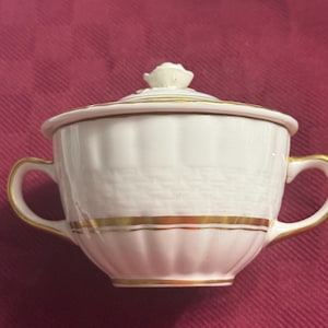 May include: White porcelain sugar bowl with a lid and two handles. The bowl has a textured surface and is accented with gold trim. The lid is topped with a small, white rose. The sugar bowl is on a red surface.