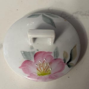May include: A white ceramic lid with a pink floral design and a small white handle. The flower has pink petals, yellow and green accents, and green leaves. The lid is likely for a teapot or similar item.