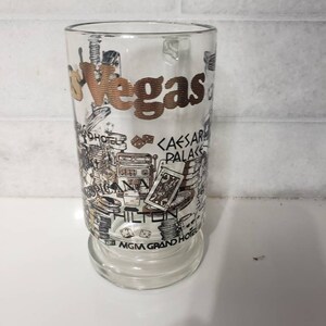 May include: A clear glass with a gold and black design featuring the word "Vegas" and various landmarks and casino names, including "Caesar's Palace", "Hilton", and "MGM Grand Hotel".