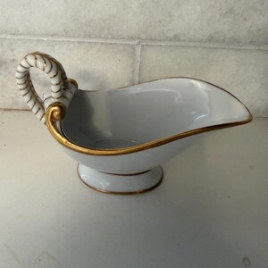 May include: A white porcelain gravy boat with a gold trim and handle. The handle has a rope-like design. The gravy boat sits on a pedestal base with gold trim. The interior is white.