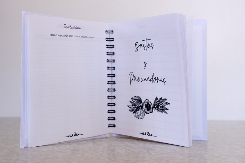 May include: Open white spiral-bound notebook with lined pages. The left page has the word "Invitaciones" and the right page has "gastos y Proveedores" in script. A floral illustration is below the text. The notebook is open on a light surface.