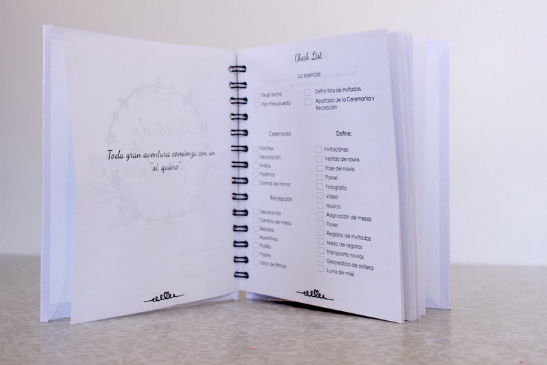May include: Open white spiral-bound notebook with checklist pages. The left page has a floral design and text in Spanish. The right page features a checklist with Spanish terms for wedding planning. The notebook is open, displaying the interior pages.