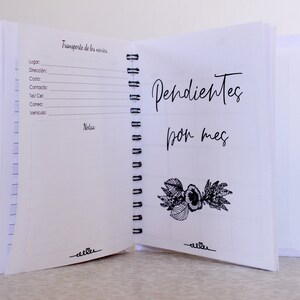 May include: Open white spiral-bound notebook with the text "Pendientes por mes" and a floral illustration. The left page has lines for notes and the text "Transporte de los novios". The notebook is open on a white surface.