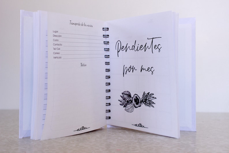 May include: Open white spiral-bound notebook with the text "Pendientes por mes" and a floral illustration. The left page has lines for notes and the text "Transporte de los novios". The notebook is open on a white surface.