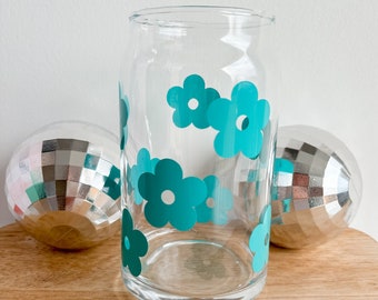 May include: A clear glass drinking glass with a teal blue daisy pattern. The glass is sitting on a wooden surface.